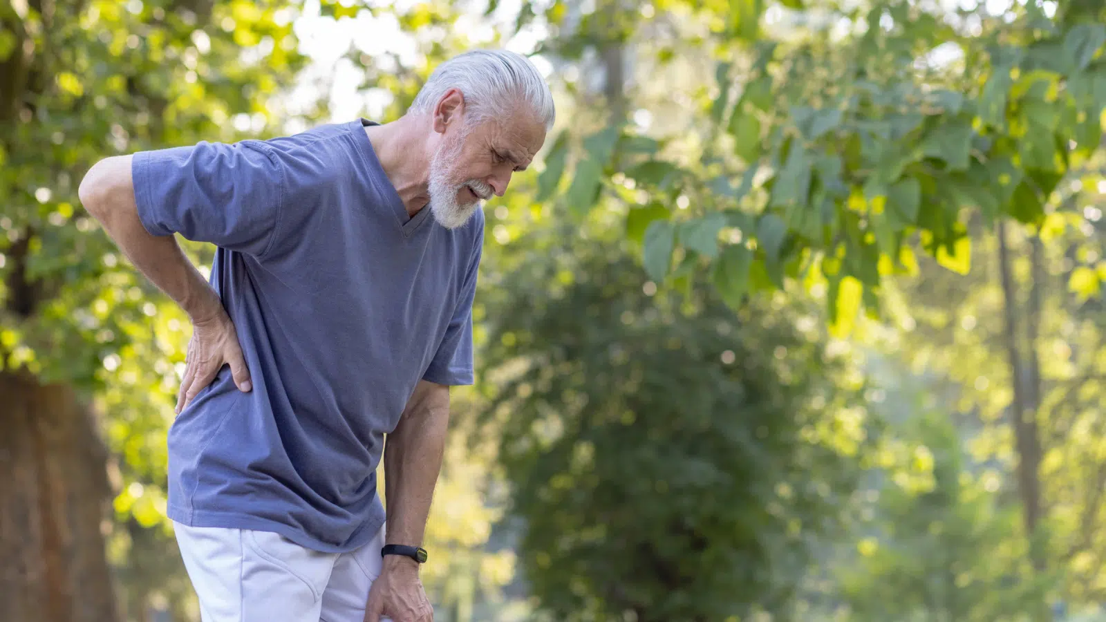 Chronic Pain Management in Older Adults Feature Image: Senior gray-haired man with a beard jogging in the park and doing sports, standing bent over and holding his hand behind his back, felt severe pain and sprain