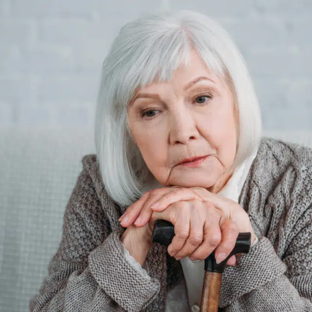Portrait of thoughtful grey hair lady with walking stick sitting on sofa at home